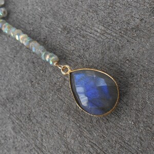 May include: A gold necklace featuring a teardrop-shaped blue labradorite gemstone pendant and a chain of iridescent beads.