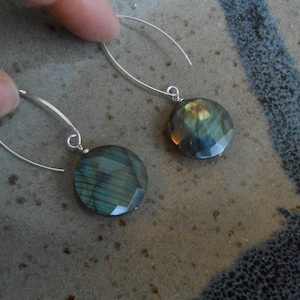 May include: A pair of silver-tone drop earrings. Each earring features a faceted, round, iridescent gemstone with shades of green, blue, and gold. The earrings are held by a person's finger.