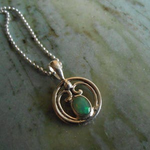 May include: Silver pendant necklace with a round, openwork design. The pendant features an oval, green gemstone. The necklace has a delicate ball chain. The jewelry is displayed on a green and gray marbled surface.