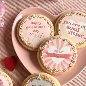 May include: Five decorated sugar cookies on a pink heart-shaped plate. The cookies have pink icing and sprinkles. The cookies have different messages on them, including "Happy Galentine's Day", "You are my soul sister", "Be my Galentine", "Fries before guys", and "Forever my best-tea".