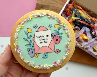 May include: A round sugar cookie with light blue icing, decorated with a pink envelope graphic containing a note that reads "Sending you a hug in the post," surrounded by colorful flowers and a border of multicolored sprinkles.