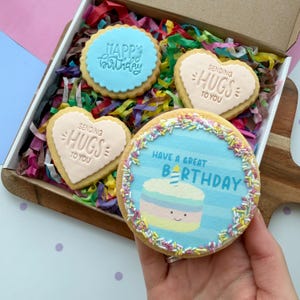 May include: A gift box containing four decorated sugar cookies nestled in colourful shredded paper. Two heart-shaped cookies are embossed with the text "SENDING HUGS TO YOU". One round cookie features "HAPPY BIRTHDAY". One large round cookie shows a cake illustration with the text "HAVE A GREAT BIRTHDAY" and rainbow sprinkles.