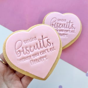 May include: Two pink heart-shaped sugar cookies with the text "sending biscuits, because you can't eat flowers" on one cookie and "sending biscuits, because you can't eat flowers" on the other cookie.