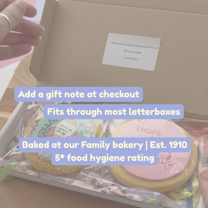 May include: A cardboard gift box containing two round cookies wrapped in clear plastic. One cookie features a floral illustration with an envelope, while the other has a pink icing disc embossed with the text I HOPE YOU SMILE.