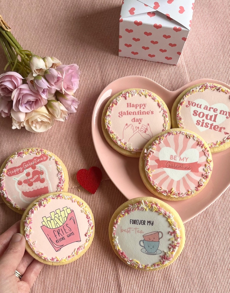 May include: Five decorated sugar cookies on a pink heart-shaped plate. The cookies have pink icing and sprinkles. The cookies have different messages on them, including "Happy Galentine's Day", "You are my soul sister", "Be my Galentine", "Fries before guys", and "Forever my best-tea".