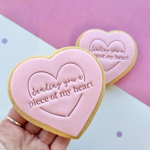 May include: Two pink heart-shaped cookies with a pink icing design. The cookies have the text "Sending you a piece of my heart" on them.