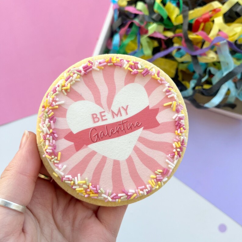 以下が含まれることがあります： ピンクと白のハート型のクッキーに「Be My Galentine」の文字が書かれています。クッキーは黄色とピンクのスプリンクルで飾られています。