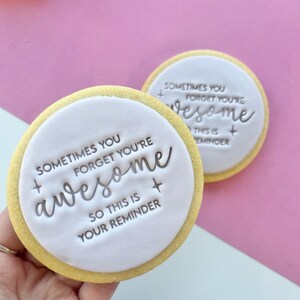 May include: Two round sugar cookies with white icing and a silver message that reads "Sometimes you forget you're awesome so this is your reminder".