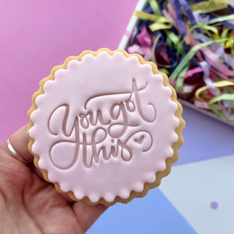 May include: A pink sugar cookie with a white scalloped edge and the words "You got this" written in cursive on top.