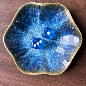 May include: A small, hexagonal ceramic dish featuring a reactive glaze with deep blue and white branching patterns, containing two translucent blue dice. The dice show white pips, with one die displaying a three and the other showing a single pip.