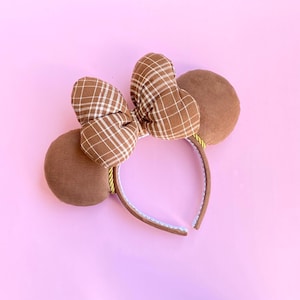 May include: A headband featuring two round brown corduroy ears and a central bow made of brown fabric with a white grid pattern. The headband frame is covered in brown fabric with a thin yellow cord trim accenting the base of the ears.