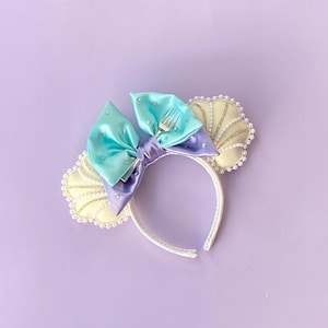 May include: A headband with a teal and purple bow, adorned with a silver fork and rhinestones. Two cream-colored seashell accents with pearl trim flank the bow. The headband is white, set against a lavender background.
