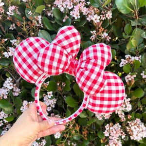 May include: A headband featuring two round ears and a large central bow, all covered in a red and white gingham checkered fabric pattern. The headband frame is wrapped in the same red and white checkered material, creating a cohesive design.