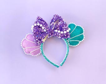 May include: Purple and teal sequined Minnie Mouse ears with a pearl and fork bow and a seashell design.