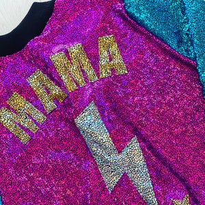 May include: A pink and blue iridescent fabric jacket with a gold "MAMA" graphic and a silver lightning bolt graphic. The jacket has a black collar and a star on the bottom right corner.
