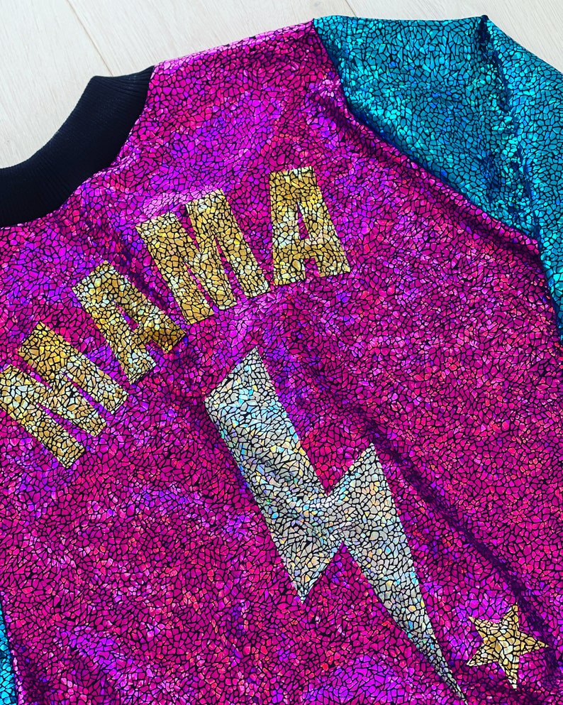 May include: A pink and blue iridescent fabric jacket with a gold "MAMA" graphic and a silver lightning bolt graphic. The jacket has a black collar and a star on the bottom right corner.