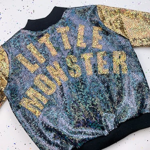 May include: A black and iridescent bomber jacket with gold lettering that reads "Little Monster".