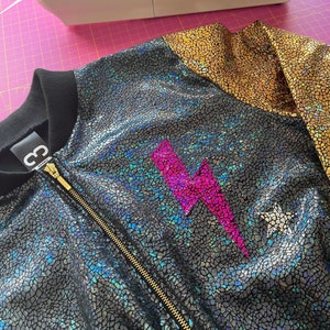 May include: A black and gold bomber jacket with a pink lightning bolt design. The jacket has a zipper closure and a ribbed collar.