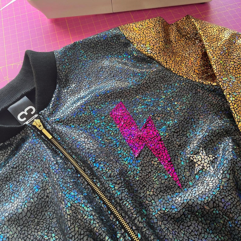 May include: A black and gold bomber jacket with a pink lightning bolt design. The jacket has a zipper closure and a ribbed collar.
