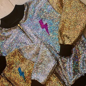 May include: A silver iridescent bomber jacket with a black zipper and black ribbed cuffs. The jacket has a lightning bolt design on the front in pink and blue.