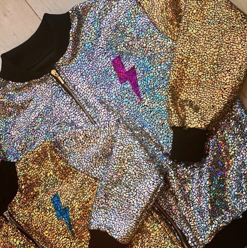 May include: A silver iridescent bomber jacket with a black zipper and black ribbed cuffs. The jacket has a lightning bolt design on the front in pink and blue.