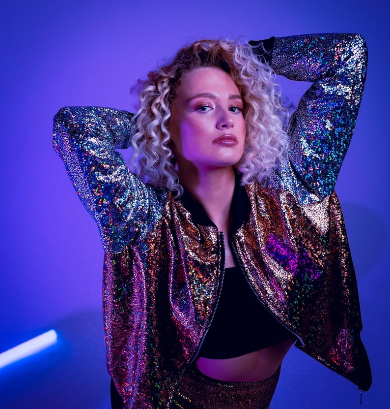 May include: A woman with curly blonde hair is wearing a iridescent bomber jacket with a black top underneath. The jacket has a zipper closure and is made of a shiny material. The woman is posing with her arms raised above her head.