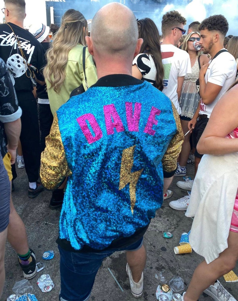 May include: A blue sequined jacket with gold sequins on the sleeves. The jacket has a pink lightning bolt and the name "DAVE" written on the back.