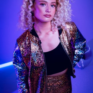 May include: A woman with curly blonde hair wears a sequined bomber jacket and matching pants. The jacket is iridescent with shades of purple, blue, and gold. The pants are a solid gold color.