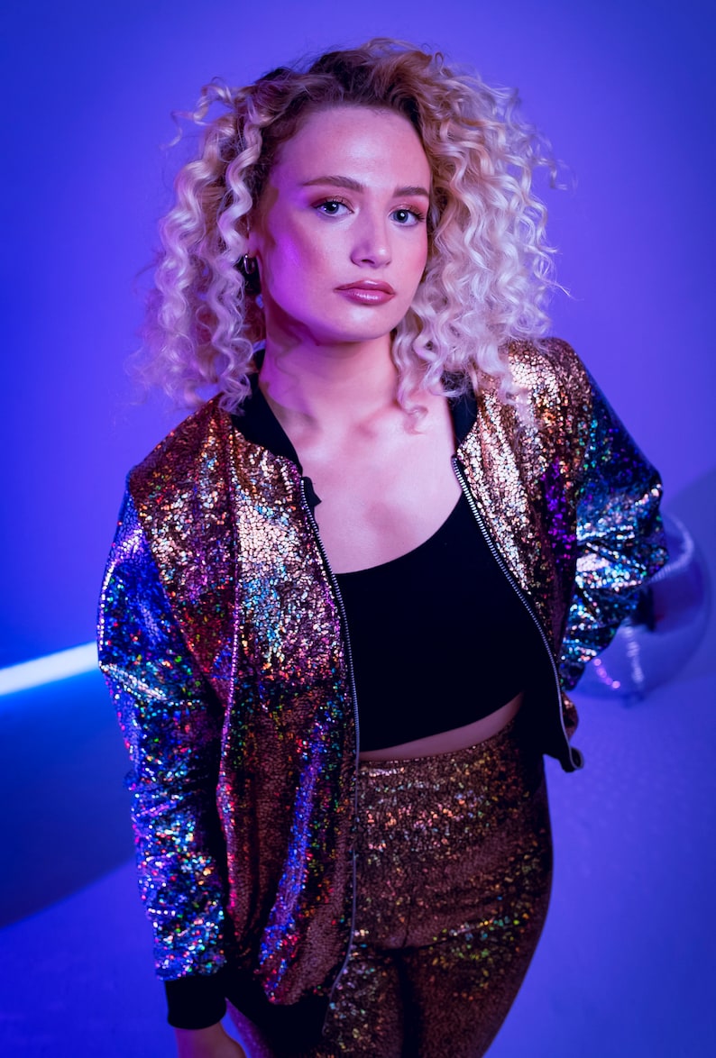 May include: A woman with curly blonde hair wears a sequined bomber jacket and matching pants. The jacket is iridescent with shades of purple, blue, and gold. The pants are a solid gold color.