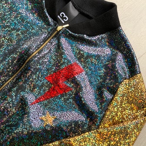 May include: A black, gold, and iridescent bomber jacket with a red lightning bolt and a star on the front. The jacket has a zipper closure and a black ribbed collar.