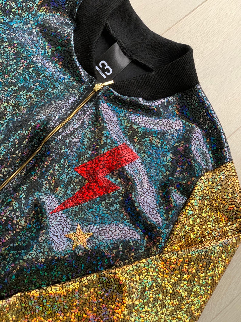 May include: A black, gold, and iridescent bomber jacket with a red lightning bolt and a star on the front. The jacket has a zipper closure and a black ribbed collar.