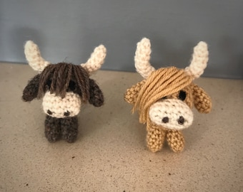 May include: Two crocheted Highland cow plushies, one brown and one tan, with white faces and fluffy yarn hair.
