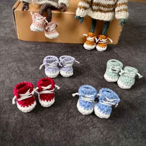 May include: A collection of miniature crochet sneakers in various colors including red, light blue, lavender, mint green, pink, and mustard yellow. Each pair features white laces and a white sole, displayed alongside two small crocheted dolls.