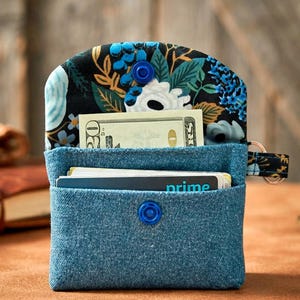 May include: A blue denim fabric wallet featuring a floral patterned lining with black, blue, and white flowers. The wallet has a blue snap closure, a main compartment containing a twenty-dollar bill, and a front pocket holding a card with the text prime.