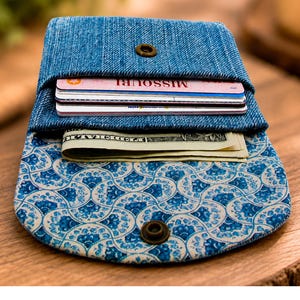 May include: A blue denim wallet featuring a flap closure with a metal snap button. The interior lining displays a blue and white scalloped pattern. The wallet contains several credit cards and a folded United States paper currency bill.