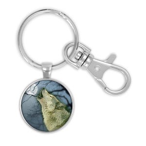 May include: Silver keychain with a large split keyring and swivel snap hook. The round pendant features a howling wolf under a full moon and bare tree branches. The text "keychain with large split keyring & swivel snap hook" is at the top.