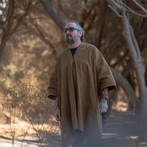 Puede incluir: Un hombre con un poncho marr&oacute;n con cremallera y una correa de cuero. Est&aacute; de pie en un bosque con &aacute;rboles en el fondo.