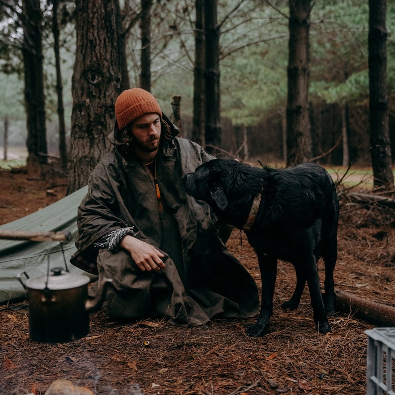 Puede incluir: Una persona con una capucha marr&oacute;n y un gorro naranja se sienta en un bosque con un perro negro. La persona sostiene una caldera de metal.