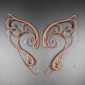 May include: A pair of copper-colored wire ear cuffs featuring intricate filigree scrollwork and curved, pointed shapes. The metal wire has a textured, beaded appearance along the outer edges, forming symmetrical, open-work designs.
