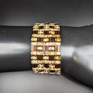 May include: A brown and gold beaded bracelet with a geometric pattern. The bracelet is made of small, square beads that are arranged in a checkered pattern.