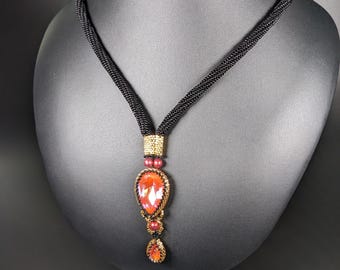 May include: A black beaded necklace with a pendant featuring a large, teardrop-shaped, orange-red crystal. The pendant is accented with gold-colored beads and smaller red crystals. The necklace is displayed on a black mannequin.