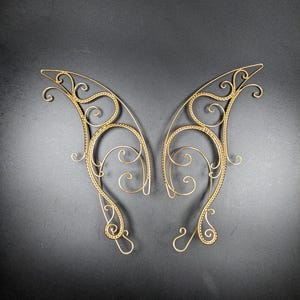 May include: A pair of gold-colored, wire-frame elf ear cuffs. The cuffs feature an intricate, swirling design with delicate curls and loops. The ear cuffs are symmetrical and appear to be made of a thin, flexible metal.
