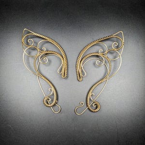 May include: A pair of gold-colored wire elf ear cuffs. The cuffs feature intricate scrollwork and beaded accents along the edges. The design includes curved lines and swirls, creating an elegant and fantasy-inspired accessory.