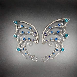 May include: A pair of silver-toned elf ear cuffs with a delicate, wire-like design. The cuffs are adorned with vibrant blue crystal beads, adding a touch of sparkle. The design features curved lines and swirls, creating an ethereal look.