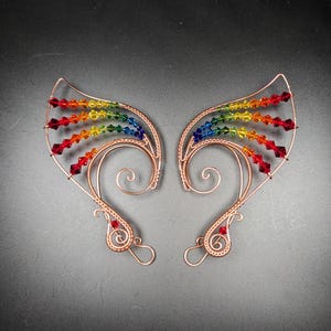 May include: A pair of copper-colored ear cuffs with a rainbow of colorful beads. The beads are arranged in a gradient of red, orange, yellow, green, blue, and purple. The ear cuffs have a delicate, wire-wrapped design with decorative swirls.