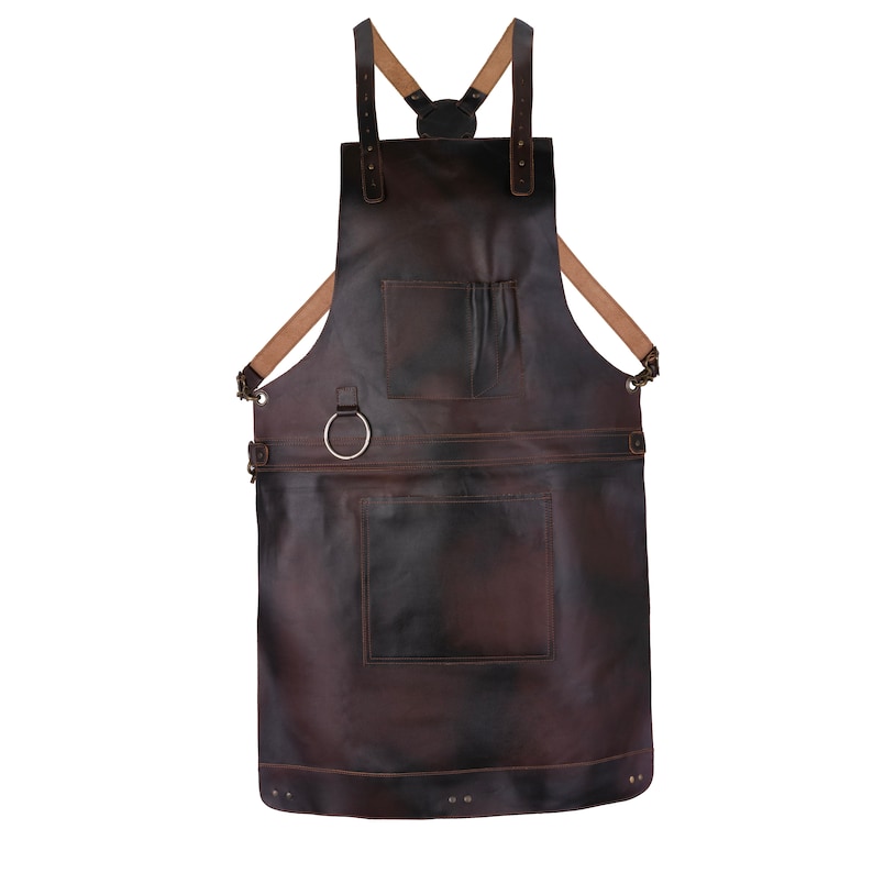 May include: A dark brown leather apron featuring adjustable tan shoulder straps that cross at the back. The apron includes a chest pocket, a large lower pocket, and a silver-toned metal ring attached to the waist area for utility.
