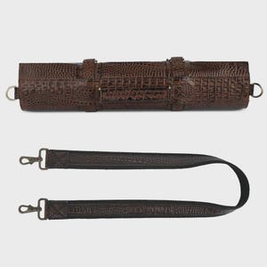 May include: A dark brown cylindrical case featuring a textured crocodile skin pattern, a central handle, and two side metal rings. It includes a detachable, adjustable shoulder strap with matching crocodile texture and metal swivel clips.