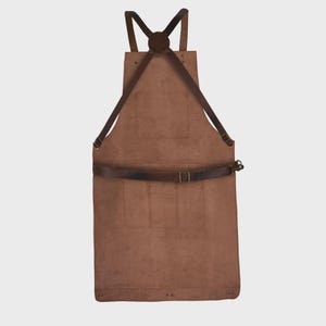 May include: A brown leather apron featuring a cross-back strap design with dark brown adjustable leather straps, a central chest pocket, and two lower pockets. The apron includes a horizontal waist strap with a metal buckle and rivets.
