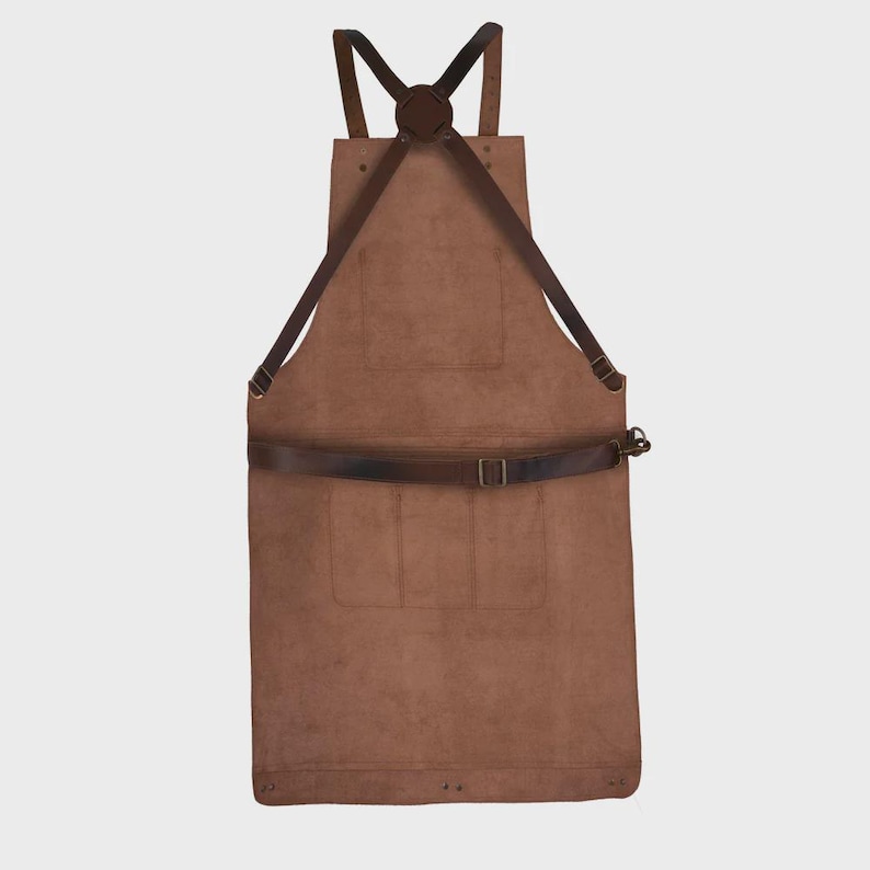 May include: A brown leather apron featuring a cross-back strap design with dark brown adjustable leather straps, a central chest pocket, and two lower pockets. The apron includes a horizontal waist strap with a metal buckle and rivets.