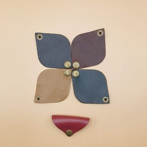May include: Five leather pieces in different colours, brown, tan, dark blue, and burgundy, are arranged in a flower shape. Each piece has a brass button in the centre. A small red leather piece with a brass button is below the flower.